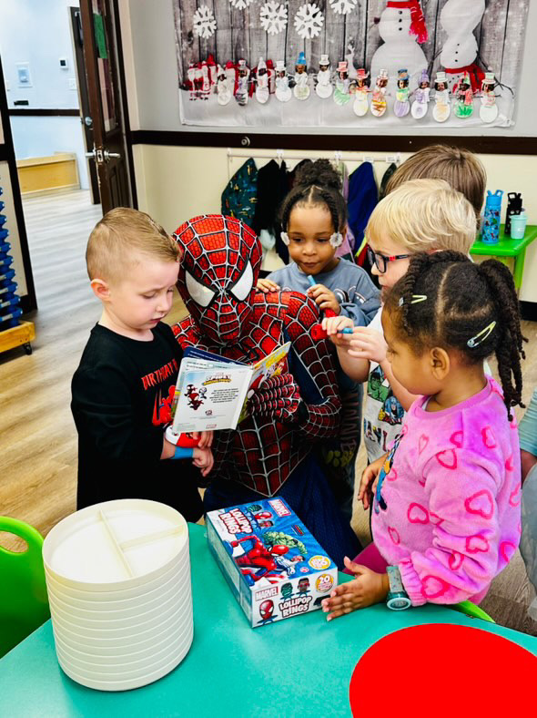 Children gather around a person dressed as Spider-Man, opening a gift in a festive setting with holiday decorations.