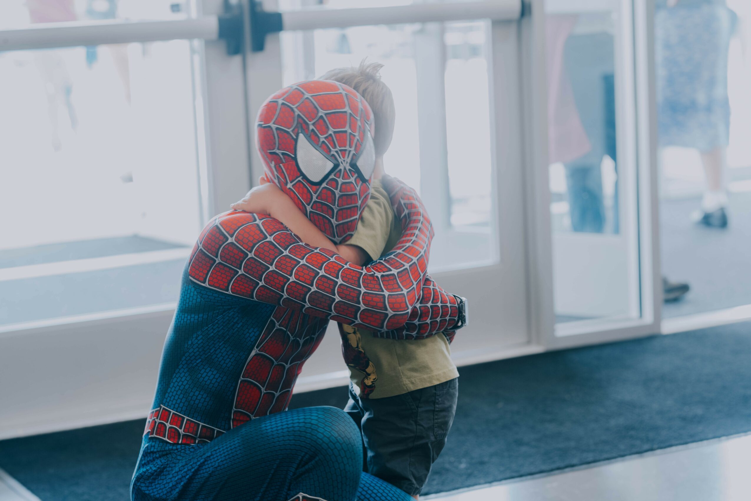 Person in Spider-Man costume hugging a child indoors near glass doors, with people visible outside.
