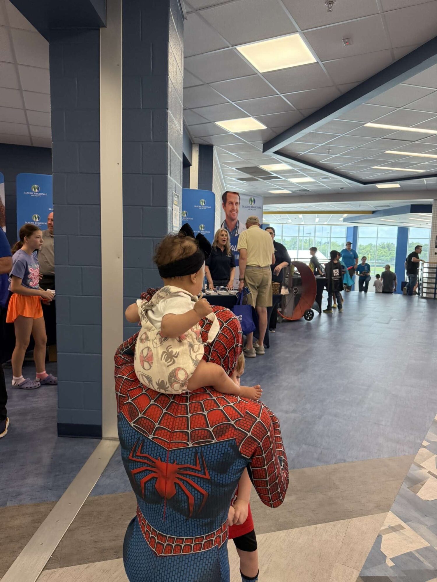 Child with a blindfold sitting on an adult's shoulders in a busy indoor public space, with people in the background.
