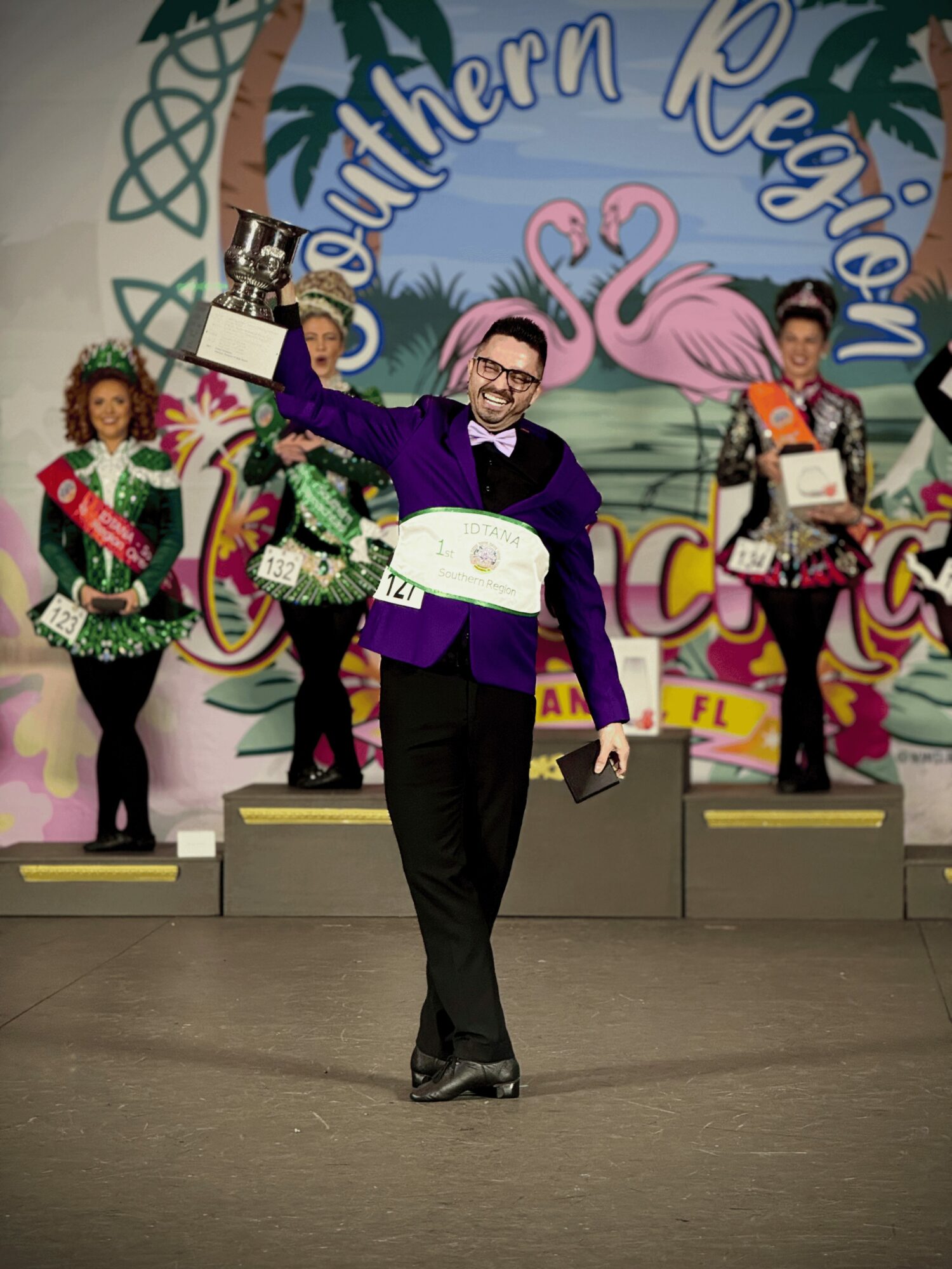 Man in purple suit holding a trophy and certificate, smiling on stage with three women in sashes and crowns, colorful background.