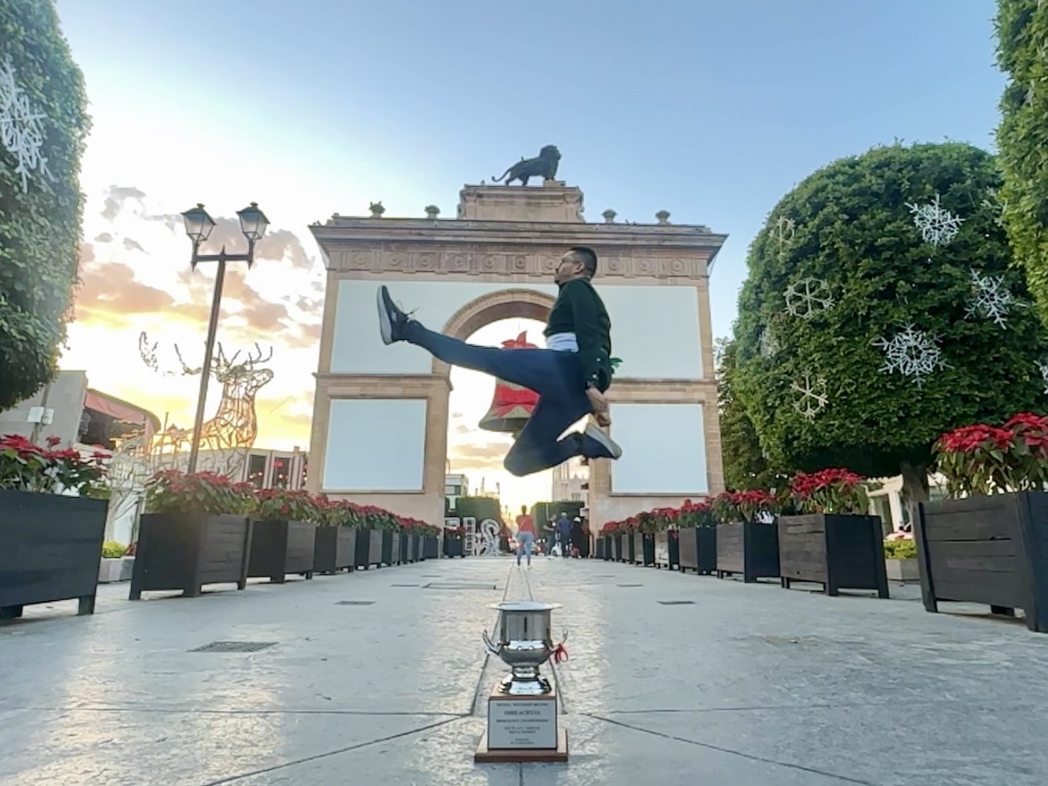 Person jumping in front of an archway, with a trophy on the ground, trees, and flower planters, during sunset.