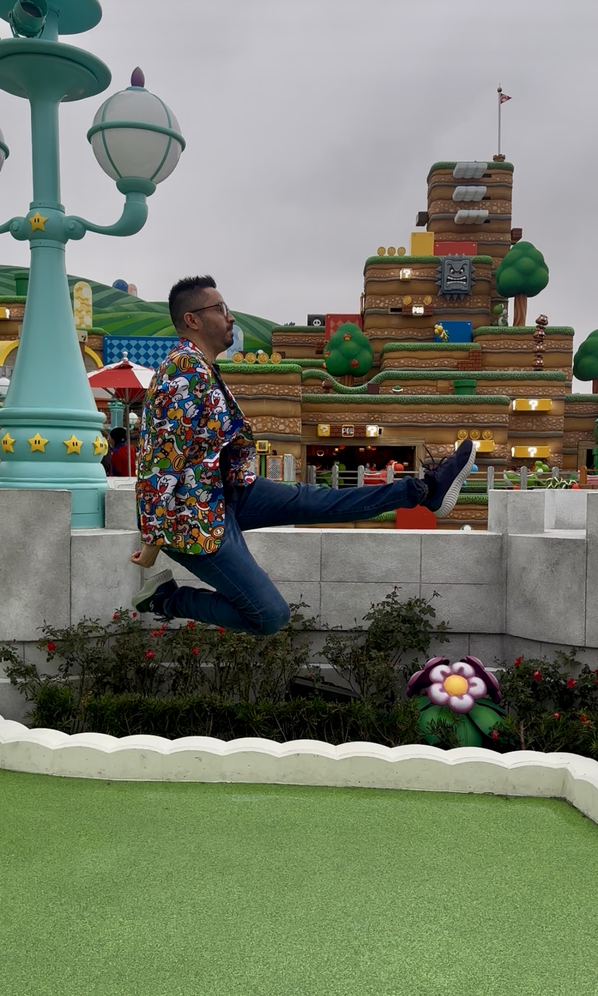 Person in colorful jacket and glasses sitting on a low wall with a theme park background, including a large building and decorative elements.