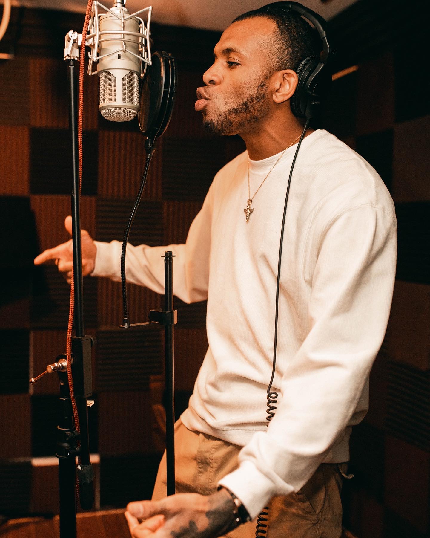 Man singing into a microphone in a recording studio, wearing a white sweatshirt and headphones, with soundproof walls.