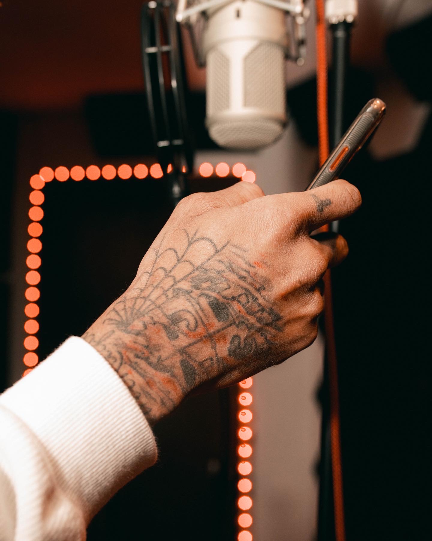 Hand with tattoos holding a smartphone, with a microphone and orange LED lights in background.