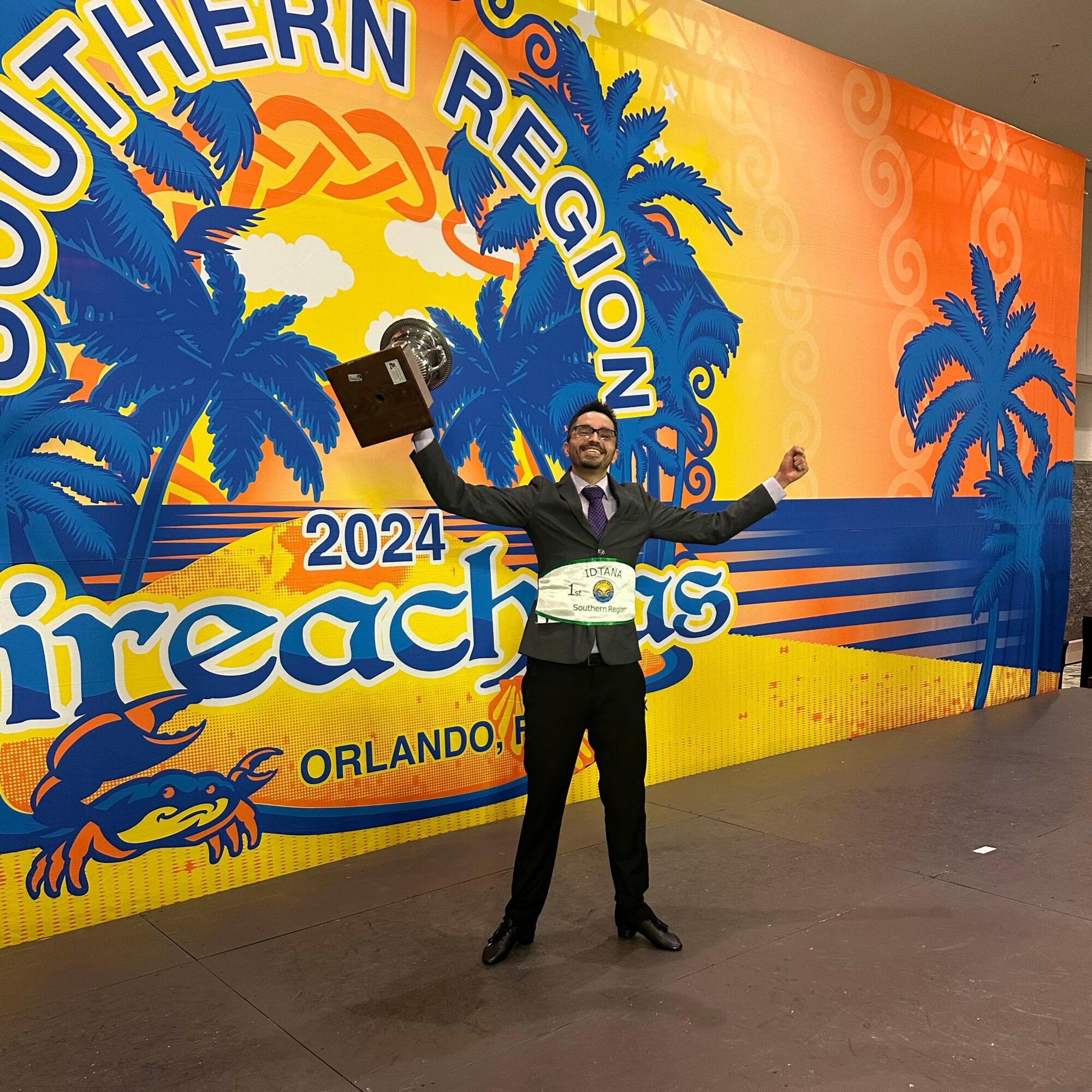 Person in suit holding a trophy in front of a colorful backdrop with palm trees and event text.