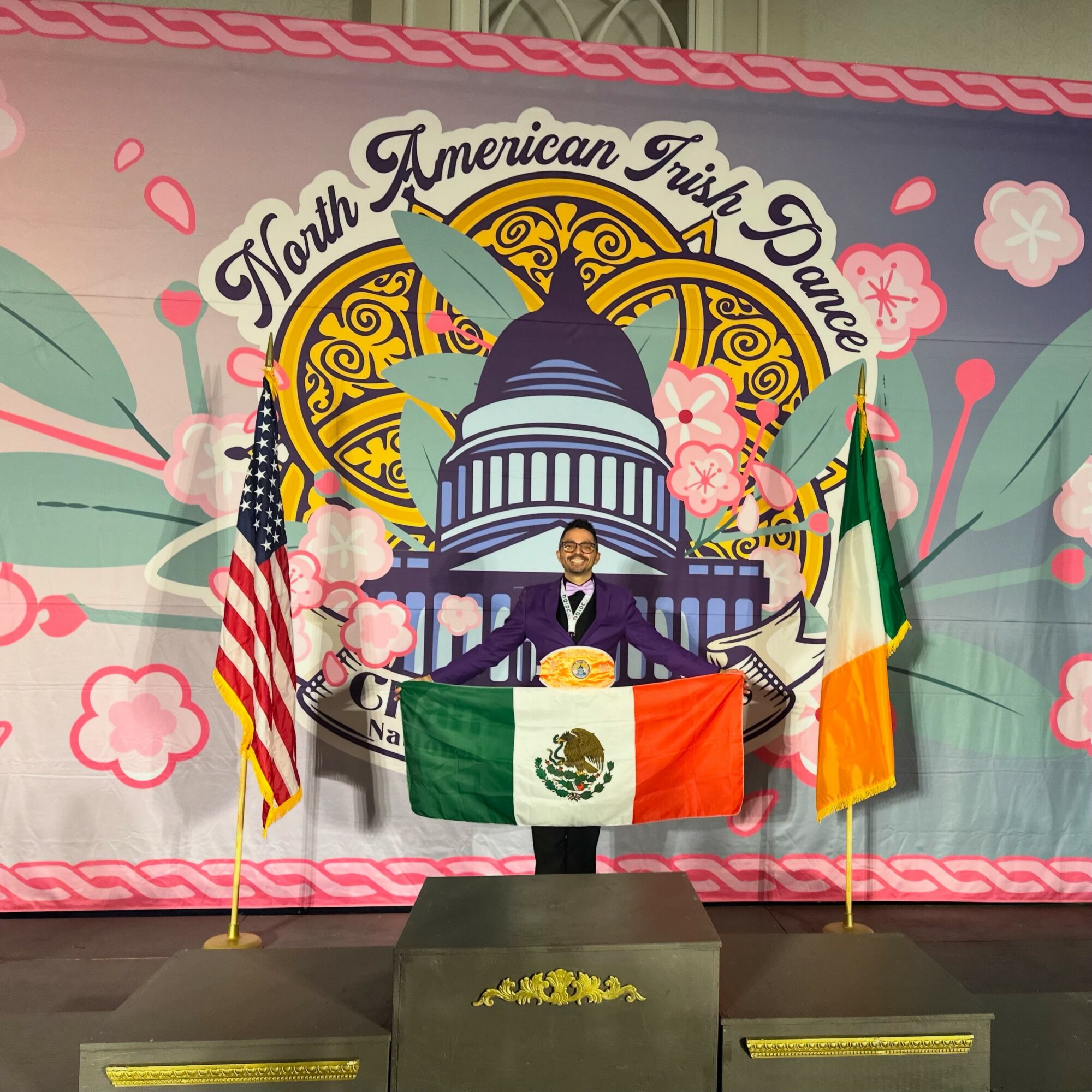 Person standing on a stage holding a Mexican flag, with American and Irish flags on either side, and a colorful backdrop with floral and architectural designs.