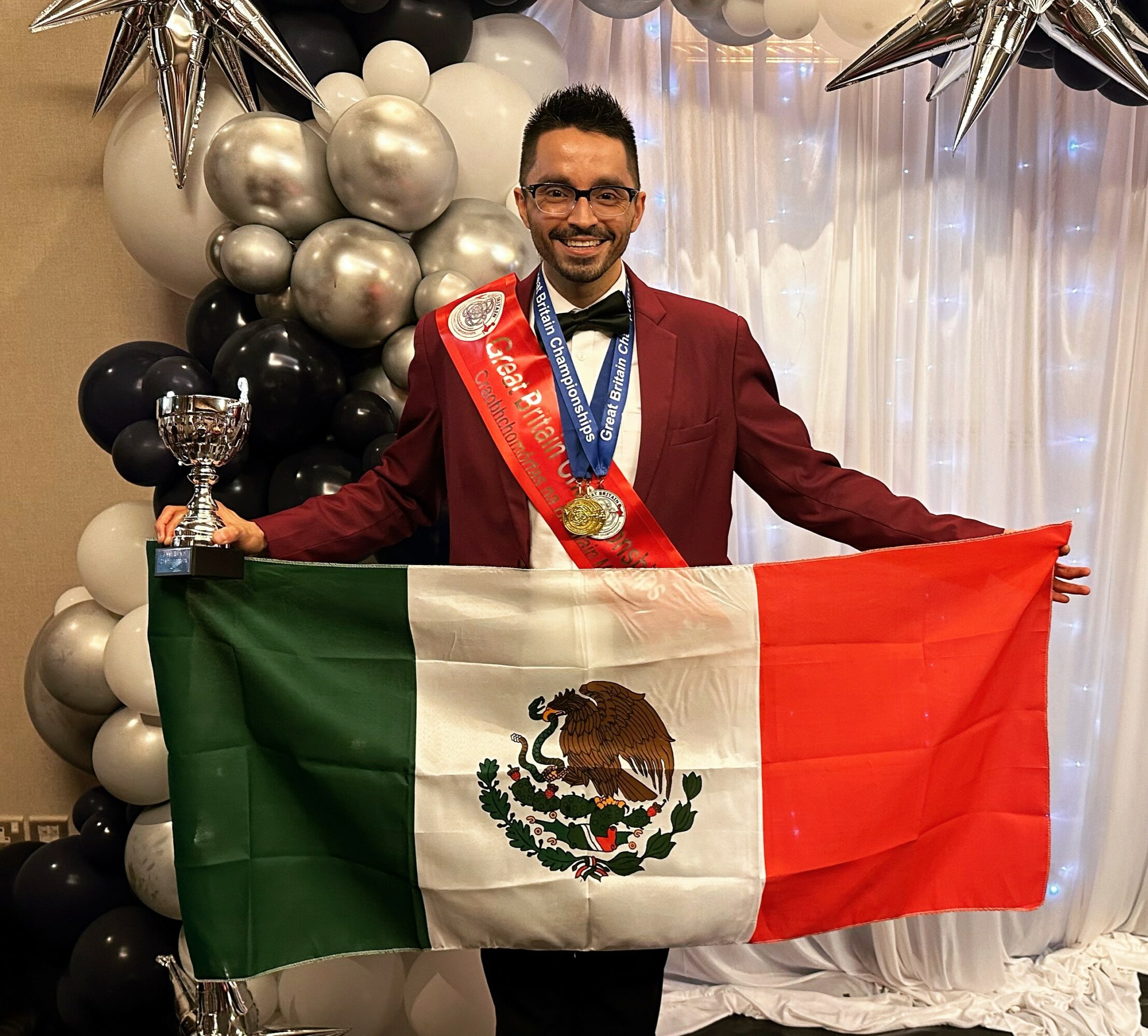 Man in formal attire holding a Mexican flag, smiling, with balloons and decorations in the background.
