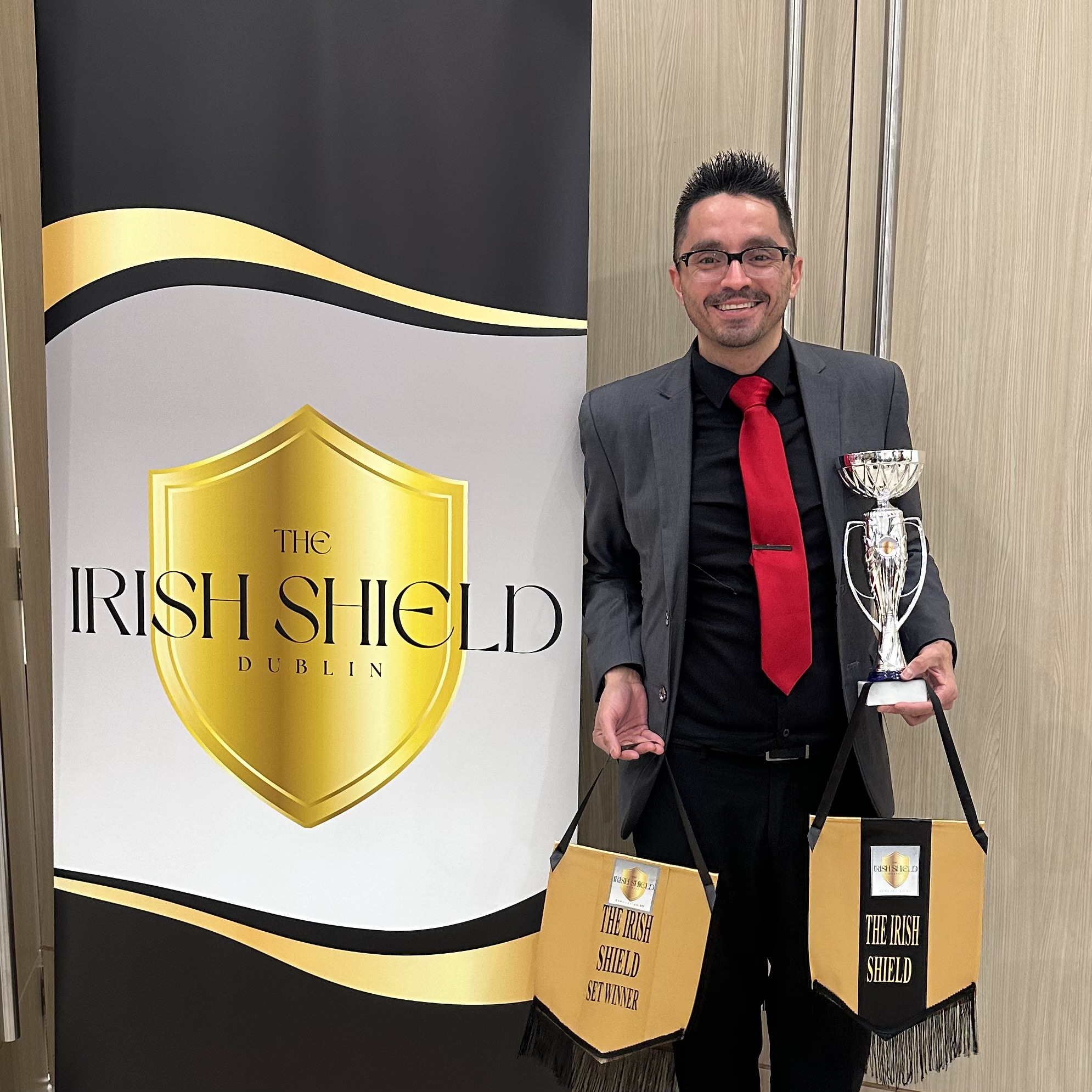 Man in suit holding a trophy and a bag, standing next to a banner with a shield logo, smiling.