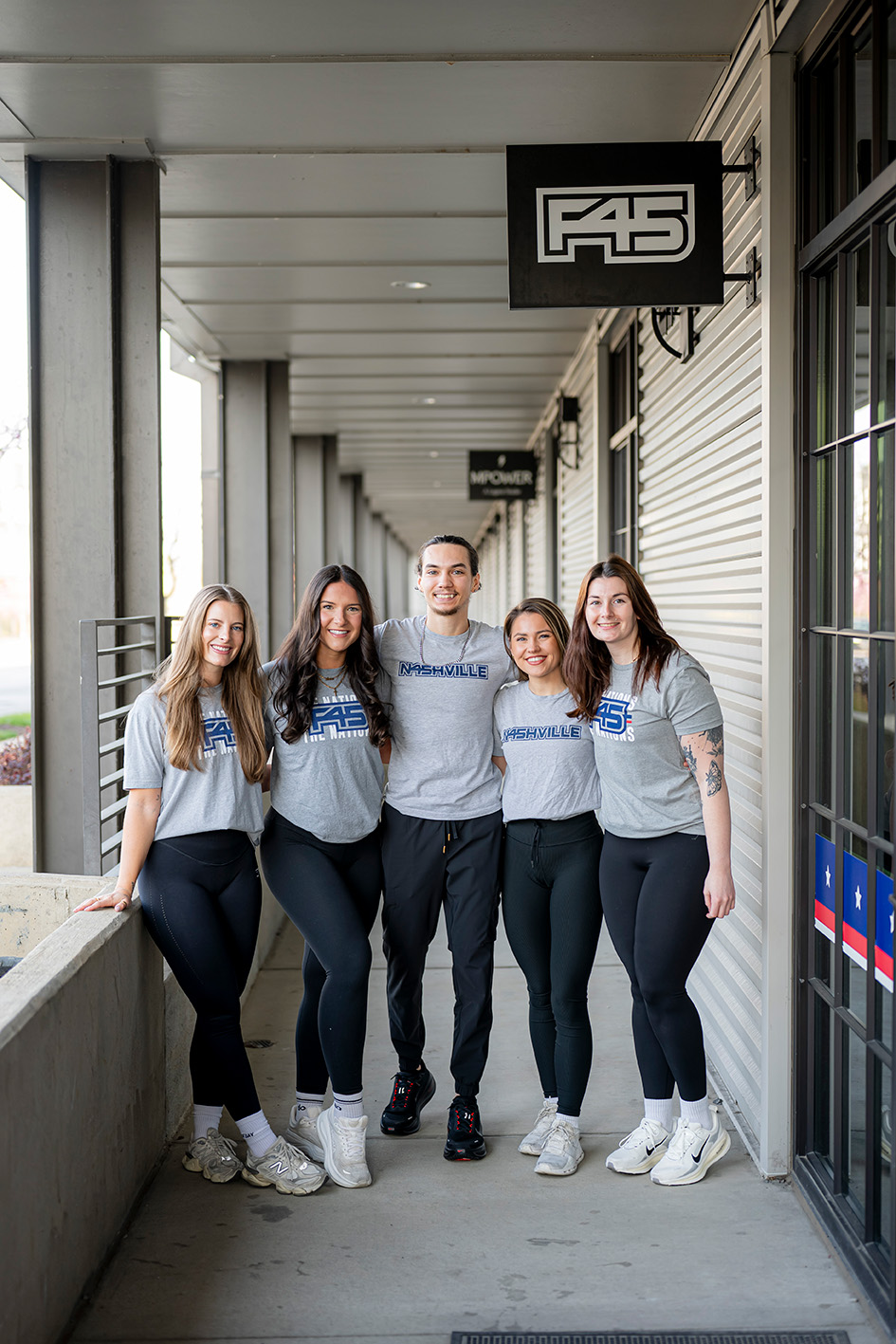 Group of five people standing in a corridor, wearing matching gray and blue shirts, smiling, with a sign above them.
