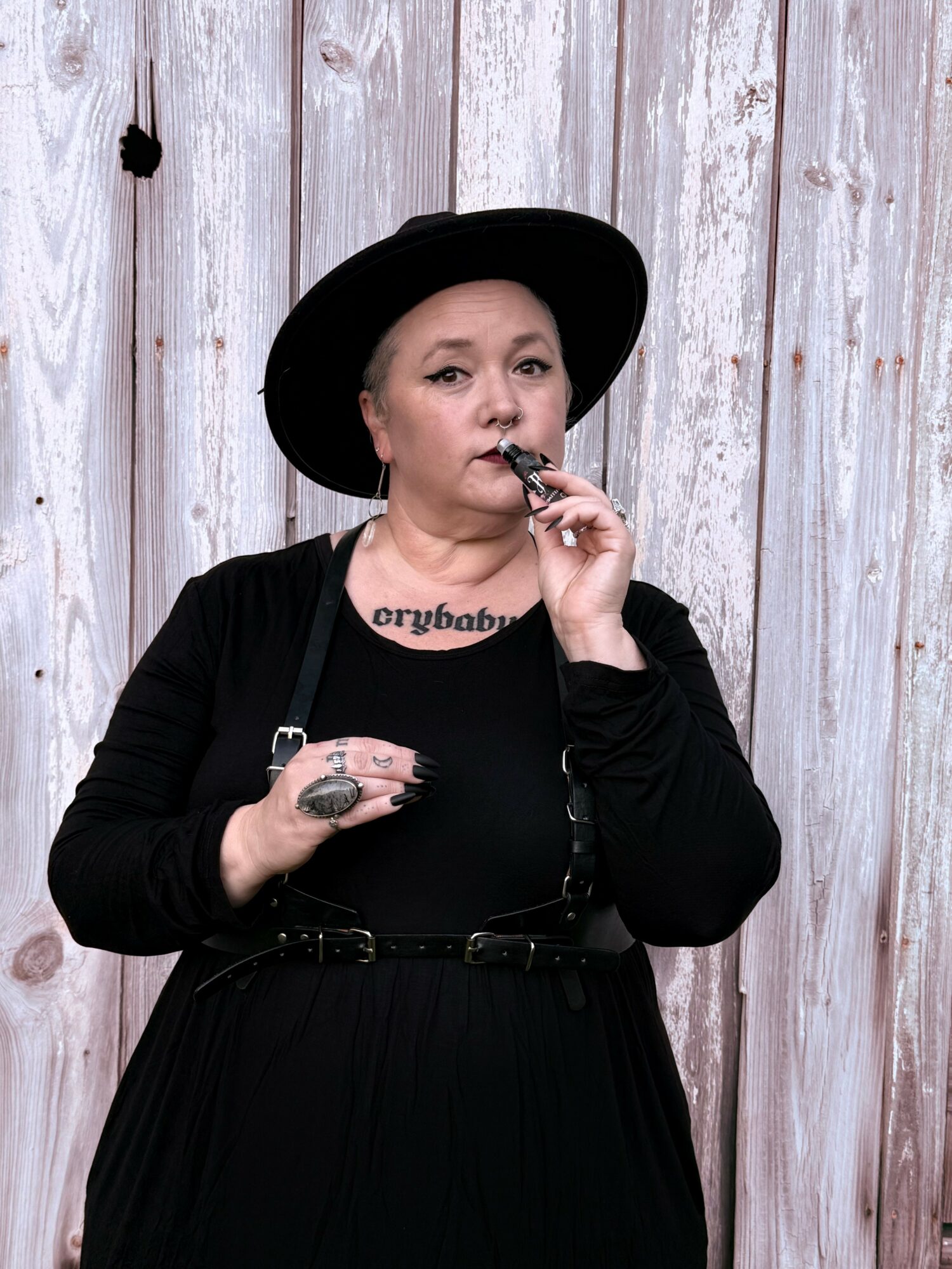 Woman with black hat and jewelry, holding a vape, standing in front of a wooden wall.