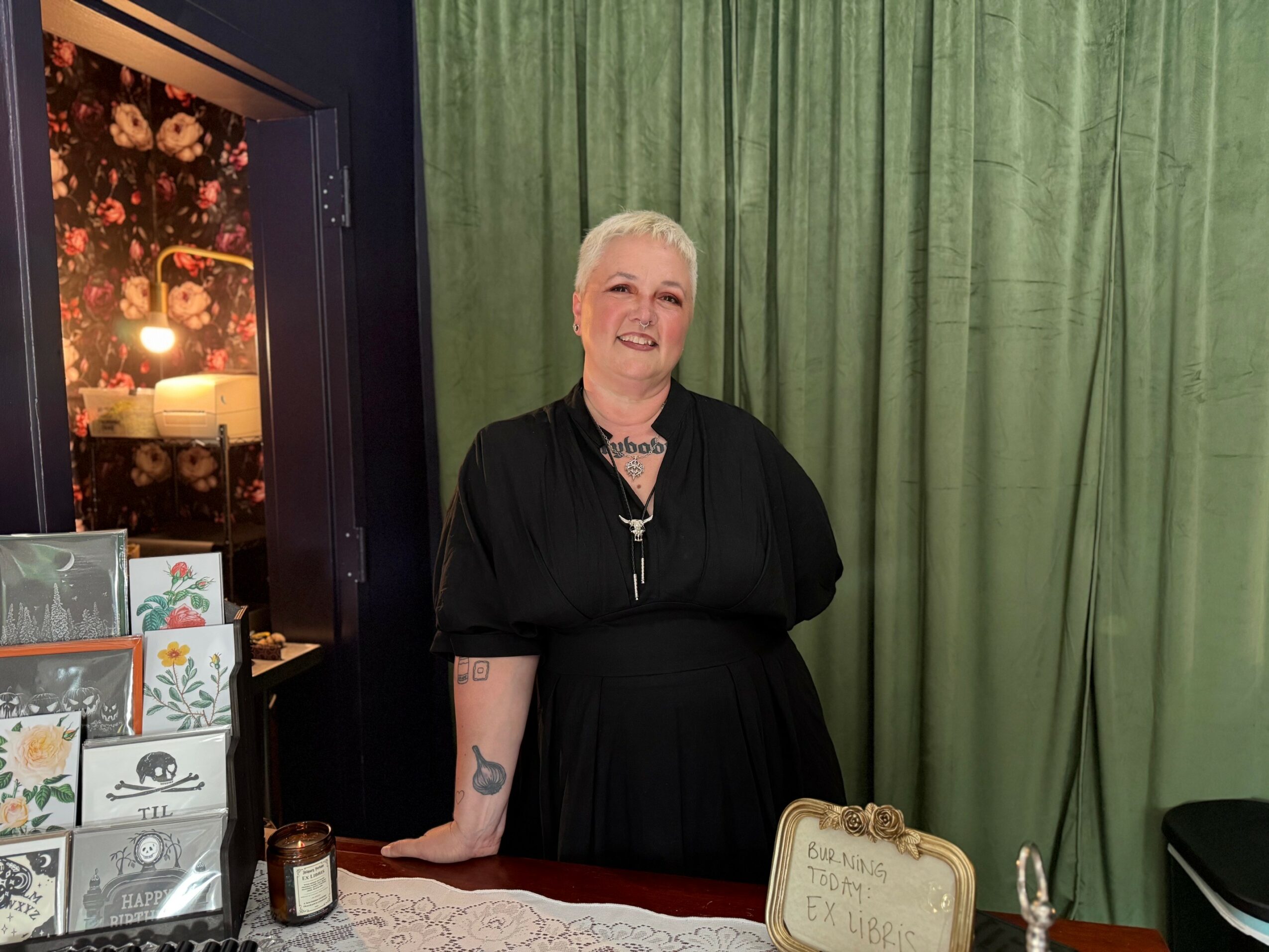 Smiling woman with short white hair standing behind a table with a green curtain background.