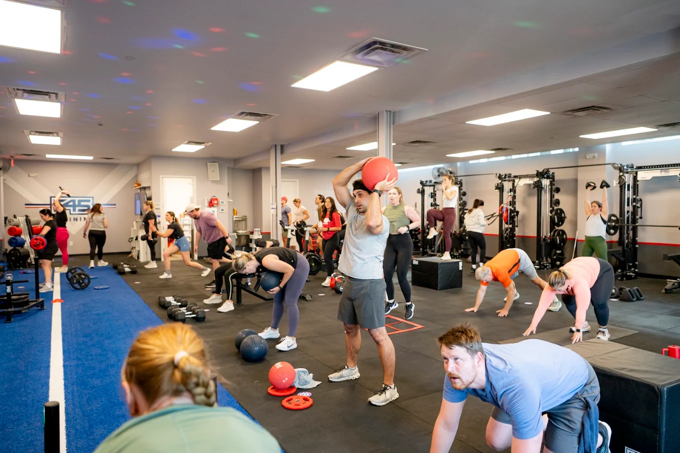 People exercising in a gym with weights and mats, some lifting kettlebells, others stretching or doing bodyweight exercises.