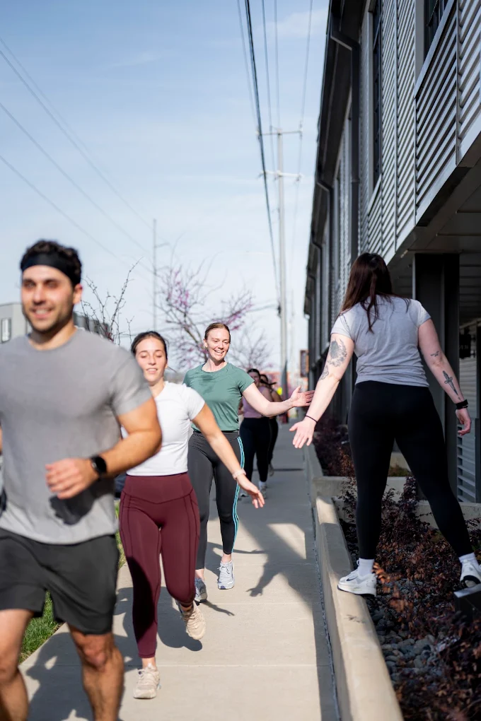 Group of people walking on sidewalk outside building, smiling and talking, sunny day, trees in background.