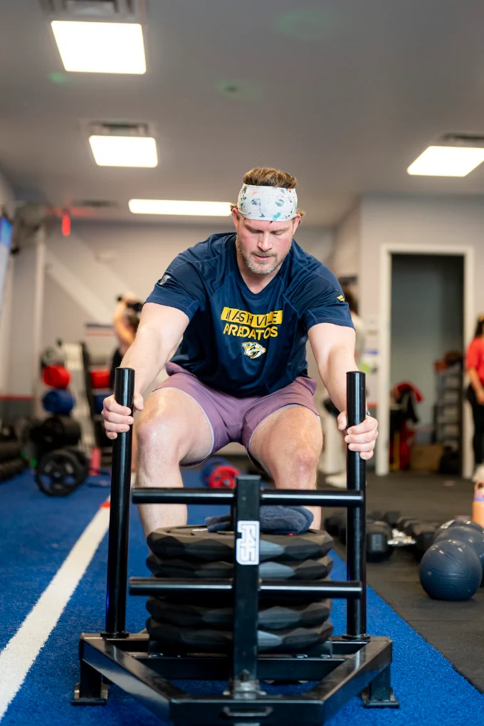 Man wearing a headband and T-shirt pushing a weighted sled in a gym, with weights and equipment in background.