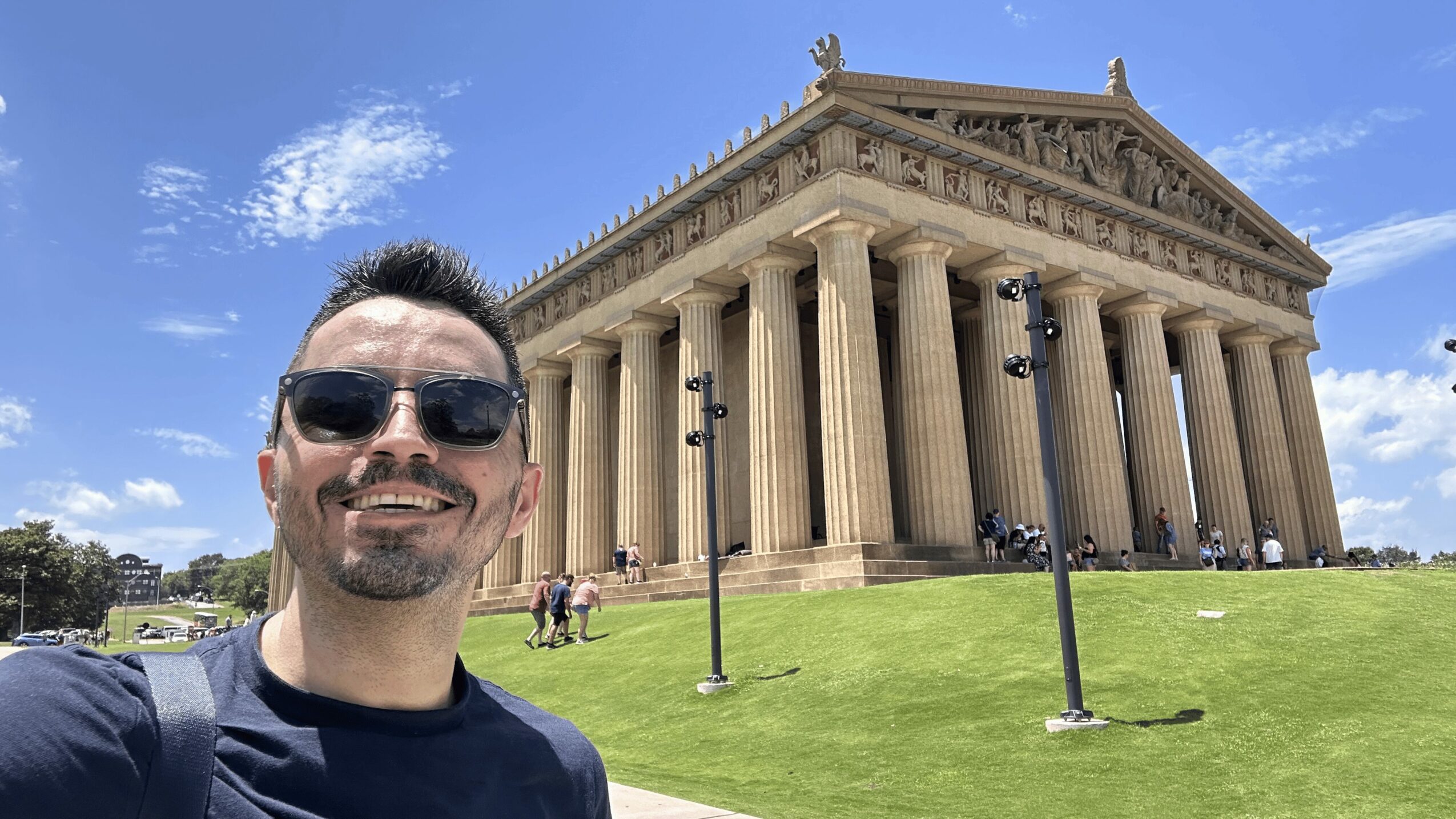 Man wearing sunglasses smiling in front of a large classical building with columns, under a blue sky.