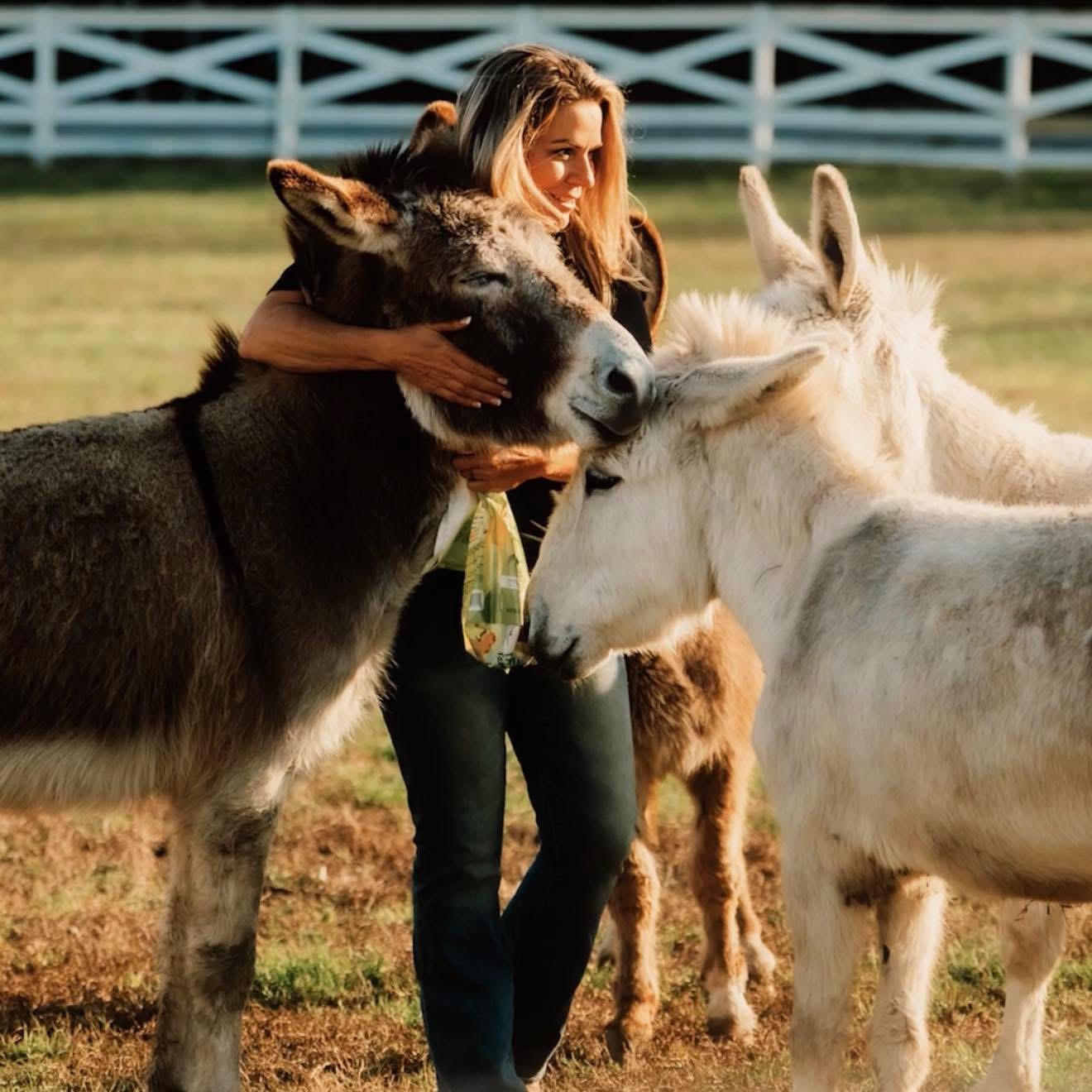 Woman hugging two donkeys in a field with a fence in the background.