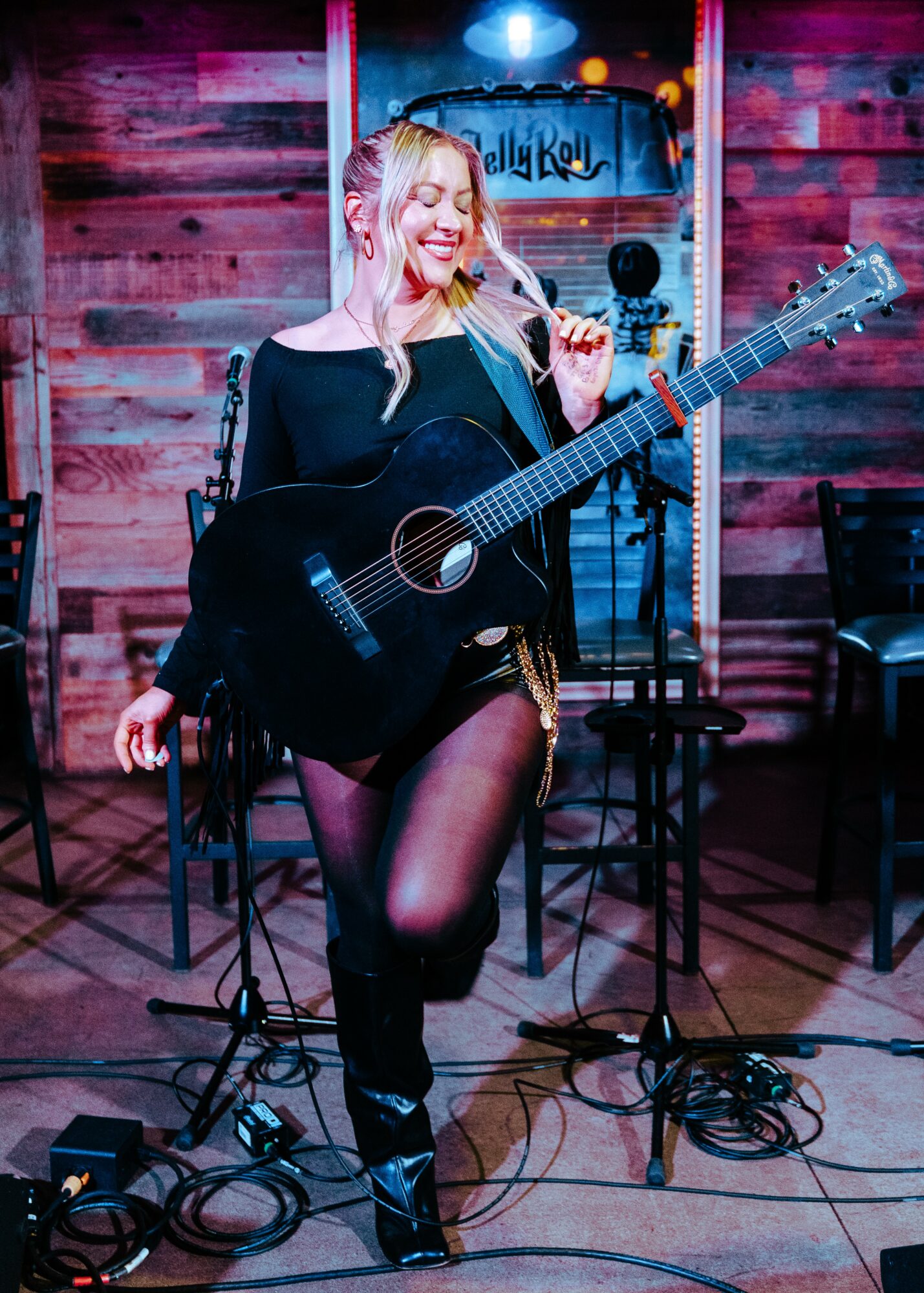 Woman with blonde hair holding a black acoustic guitar on stage, smiling, in a dimly lit venue with wooden walls.