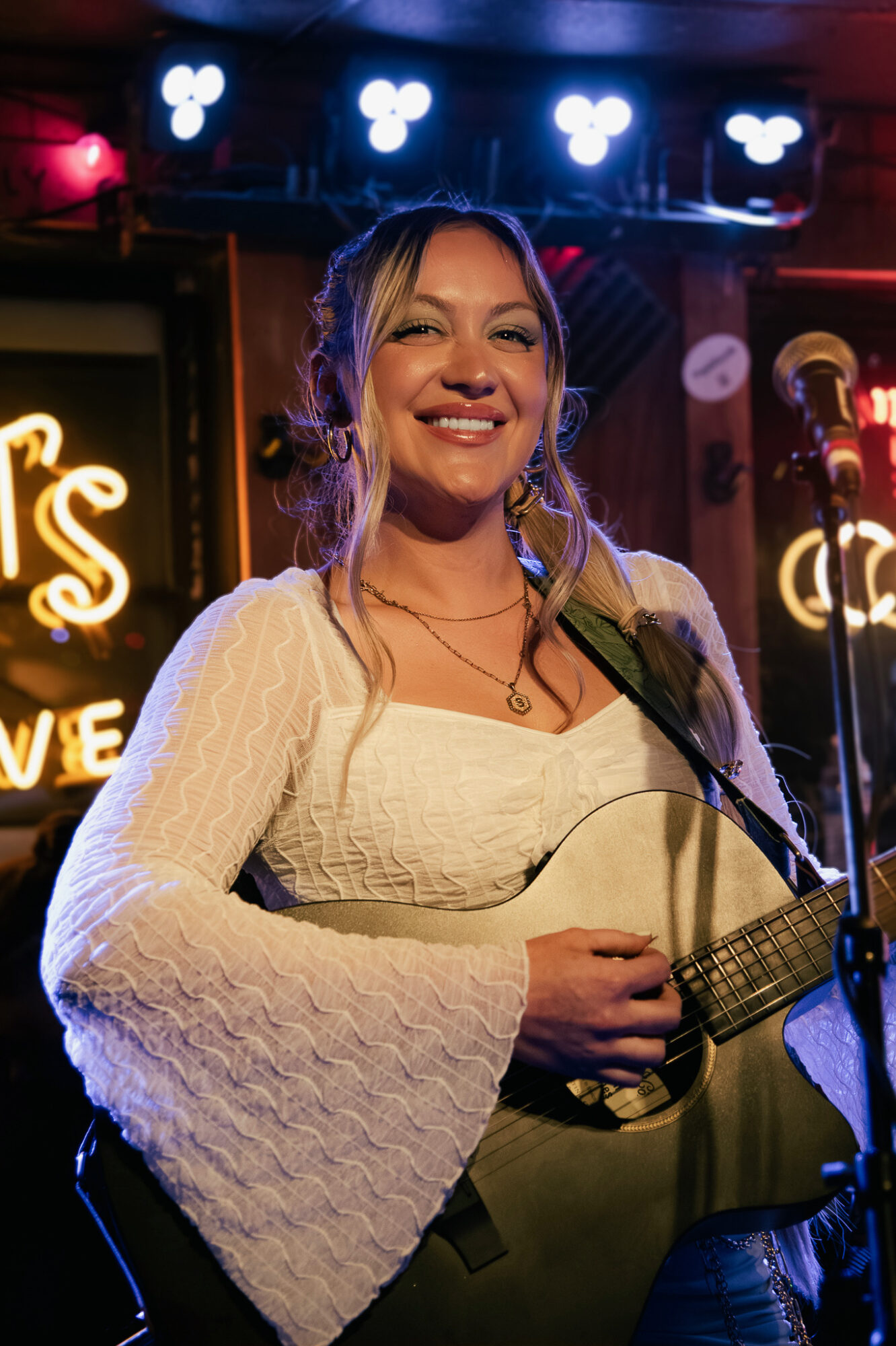 Woman smiling with a guitar on stage, microphone nearby, illuminated by colorful stage lights.