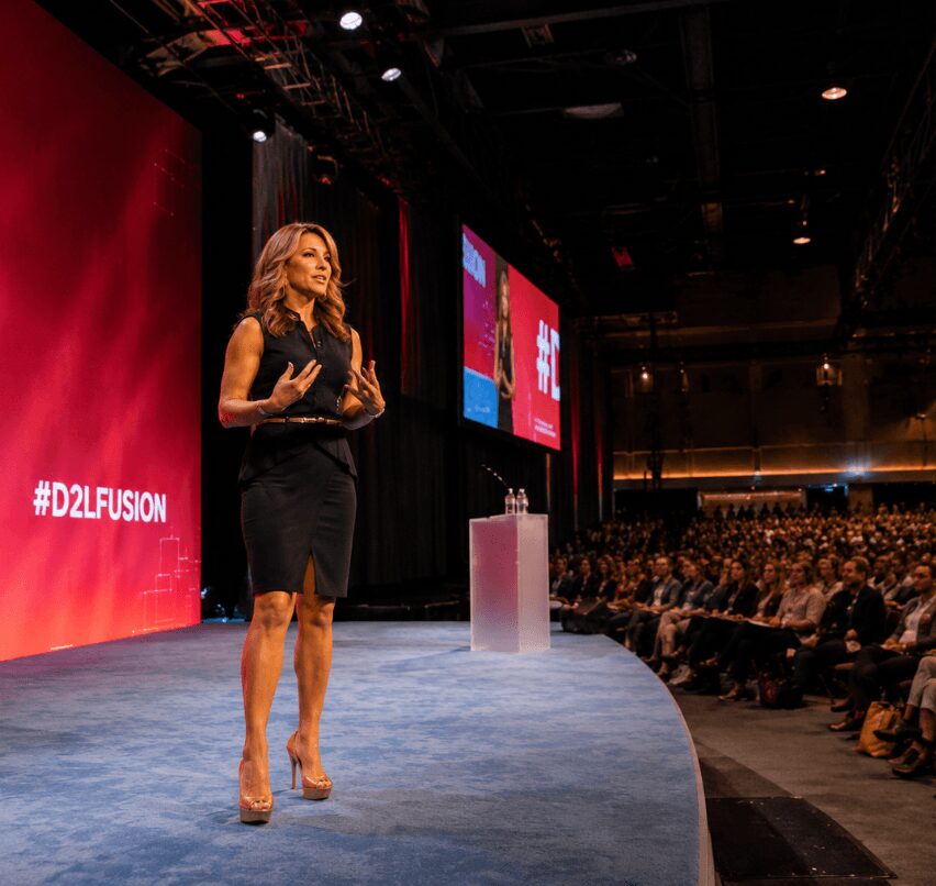 Woman speaking on stage at conference with audience in background, red backdrop, and screens displaying event graphics.