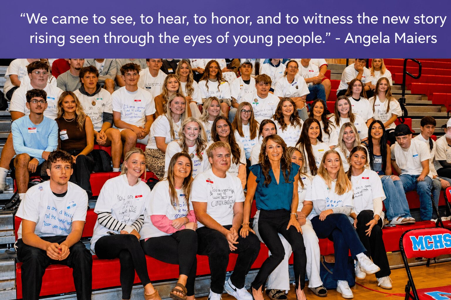 Group of children and adults sitting on red bleachers, some wearing white T-shirts, in a school or event setting.