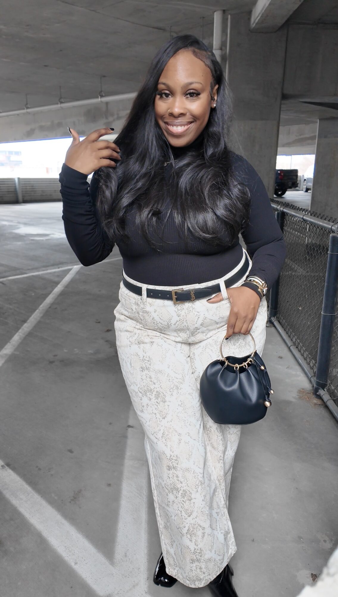 Smiling woman with long black hair in a parking garage, wearing a black top, patterned pants, and holding a small black purse.