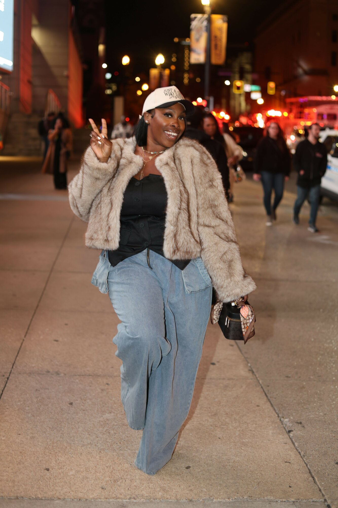 Person walking on sidewalk at night, wearing a fur coat, wide pants, and glasses, with city lights in background.