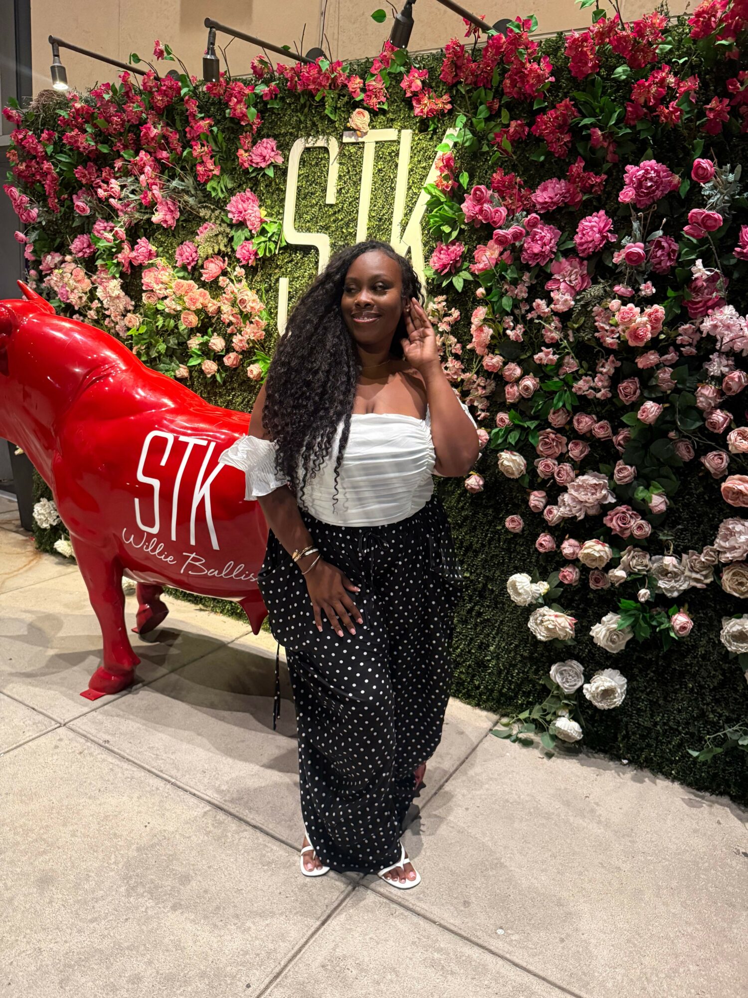 Woman with long braids in a white top and black polka dot pants standing next to a red heart-shaped sculpture with white text, flowers in background.