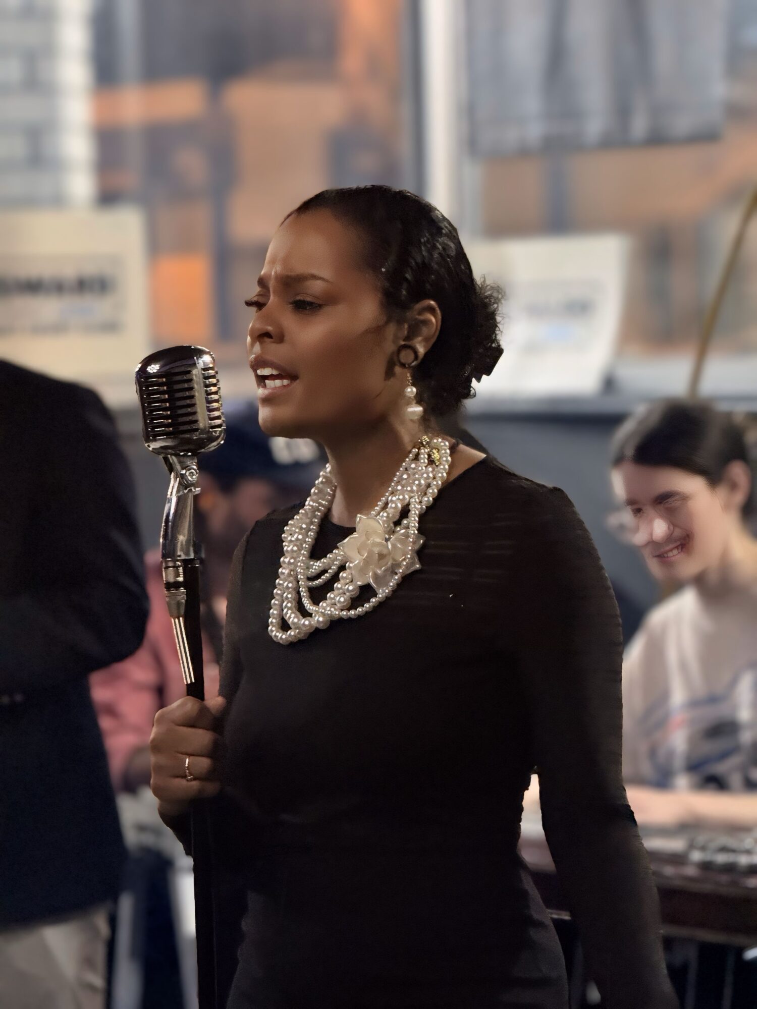 A woman singing into a vintage microphone, wearing a black dress and pearl jewelry, with a person playing keyboard in background.