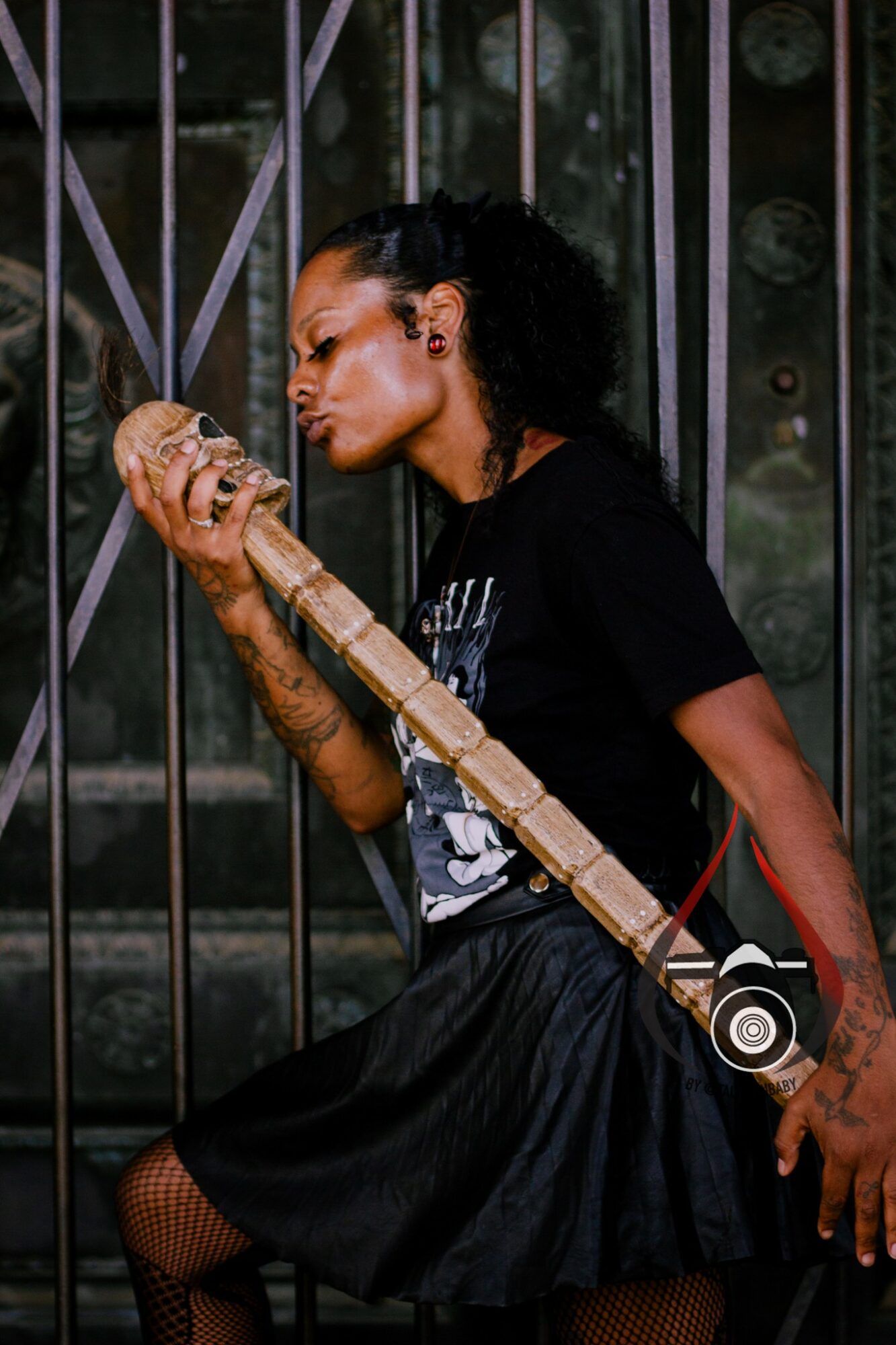 Woman with curly hair playing a long wooden flute, standing in front of metal bars, wearing a black t-shirt and skirt.