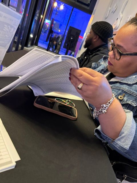 Person with glasses and bracelet writing in a notebook at a table, with a smartphone nearby, in a public space.