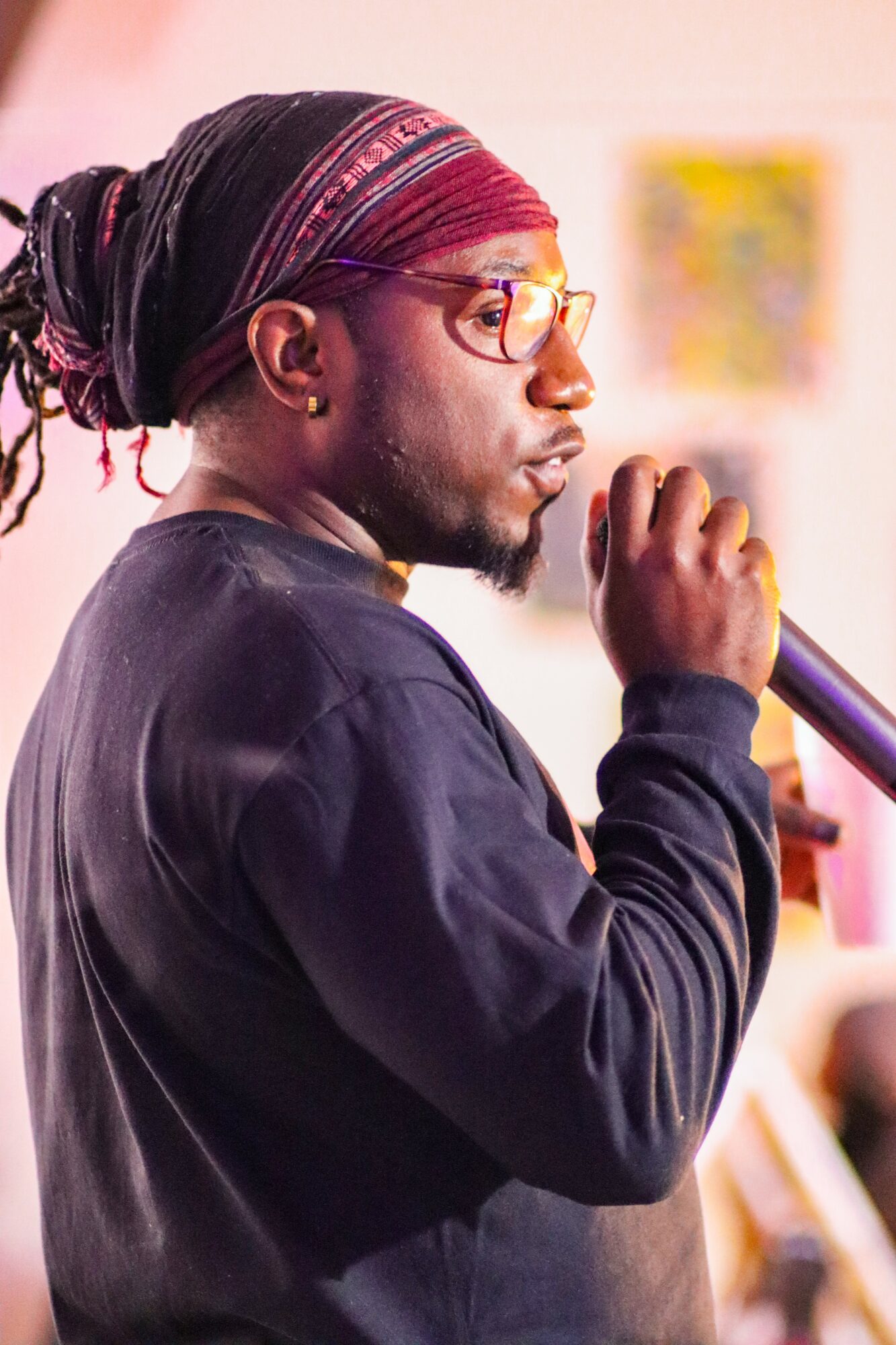 Person with glasses and headscarf holding a microphone, facing sideways, indoors, with blurred background.
