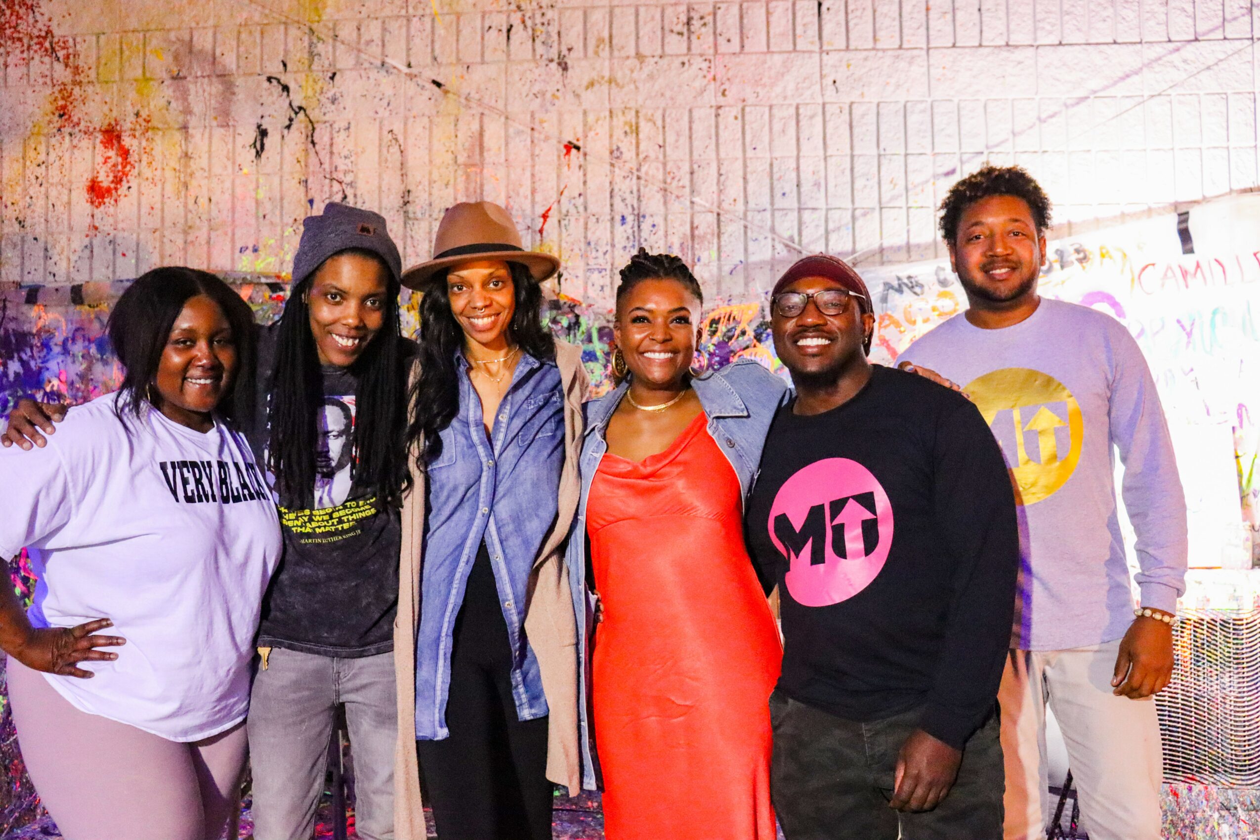 Group of six diverse people smiling, standing against a graffiti wall, wearing casual clothing.
