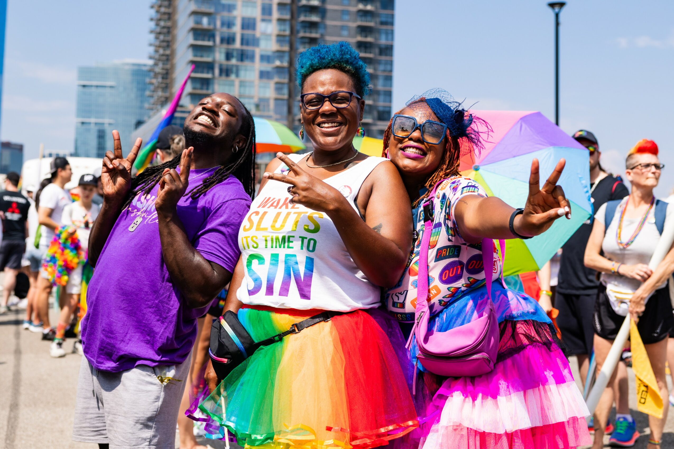 Three smiling women holding umbrellas and making peace signs at a pride parade, with city buildings in background.