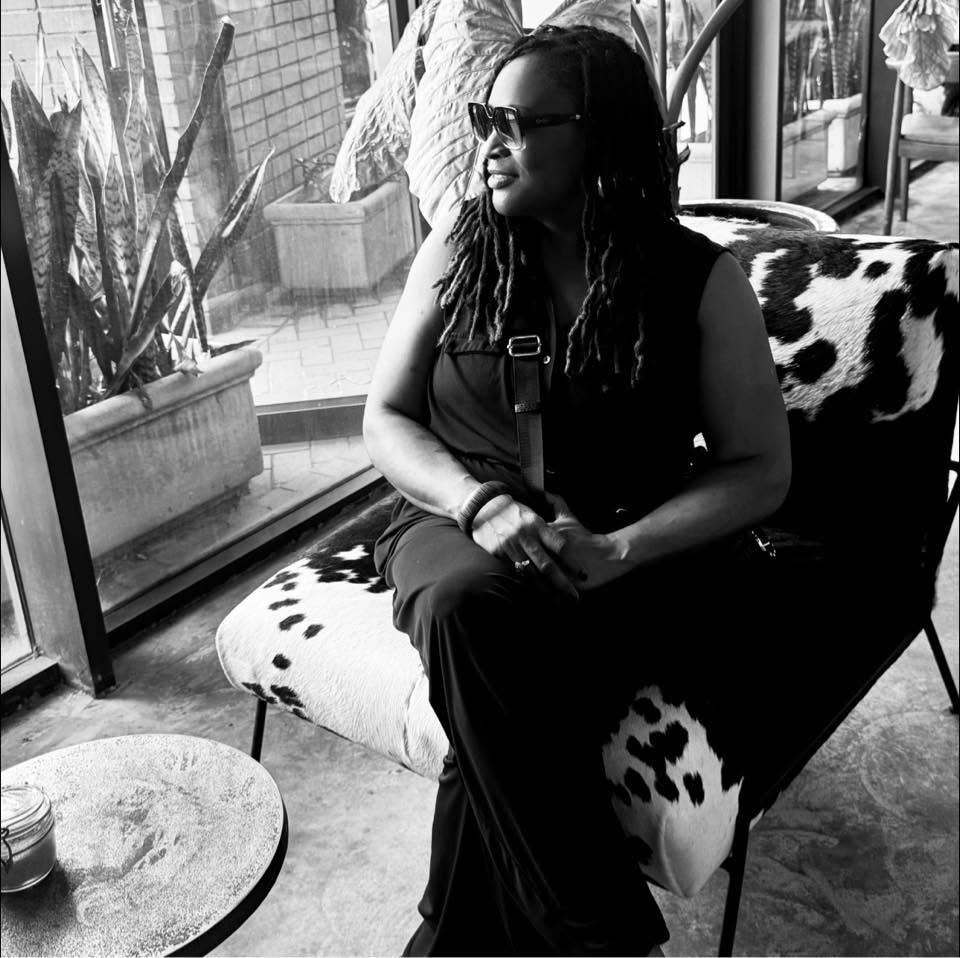 Woman with sunglasses sitting on a cow-patterned chair near a window, with plants outside, in black and white.