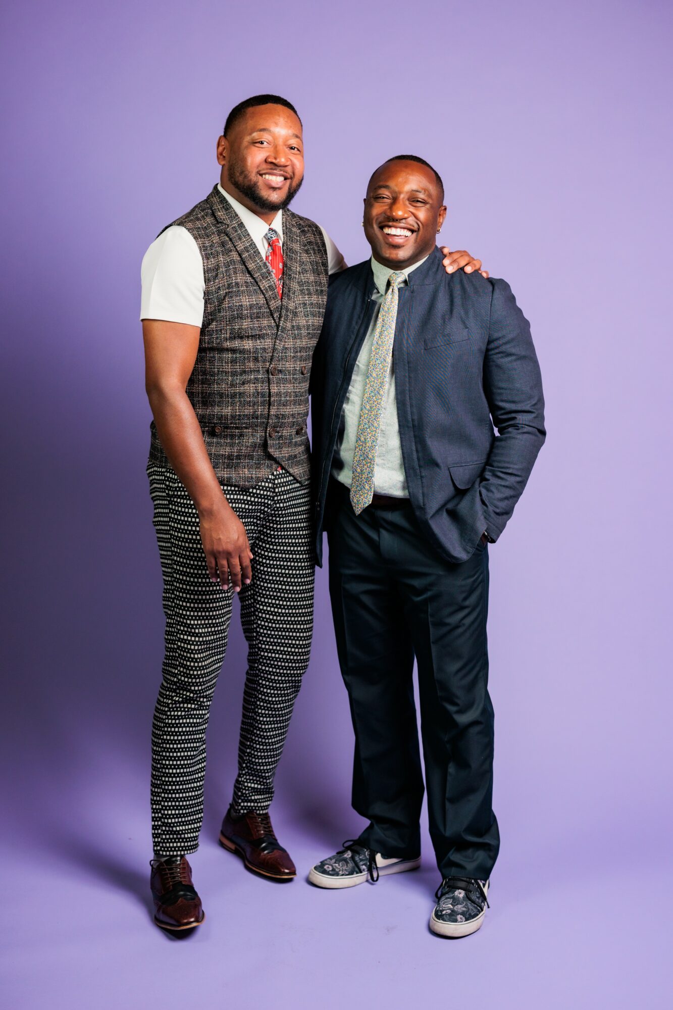 Two men stand together smiling, one with arm around the other's shoulder, against a purple background.