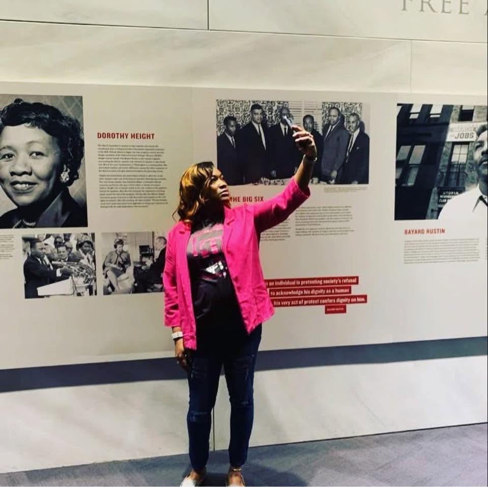 Woman in pink jacket taking a selfie in front of a display with historical photos and text.