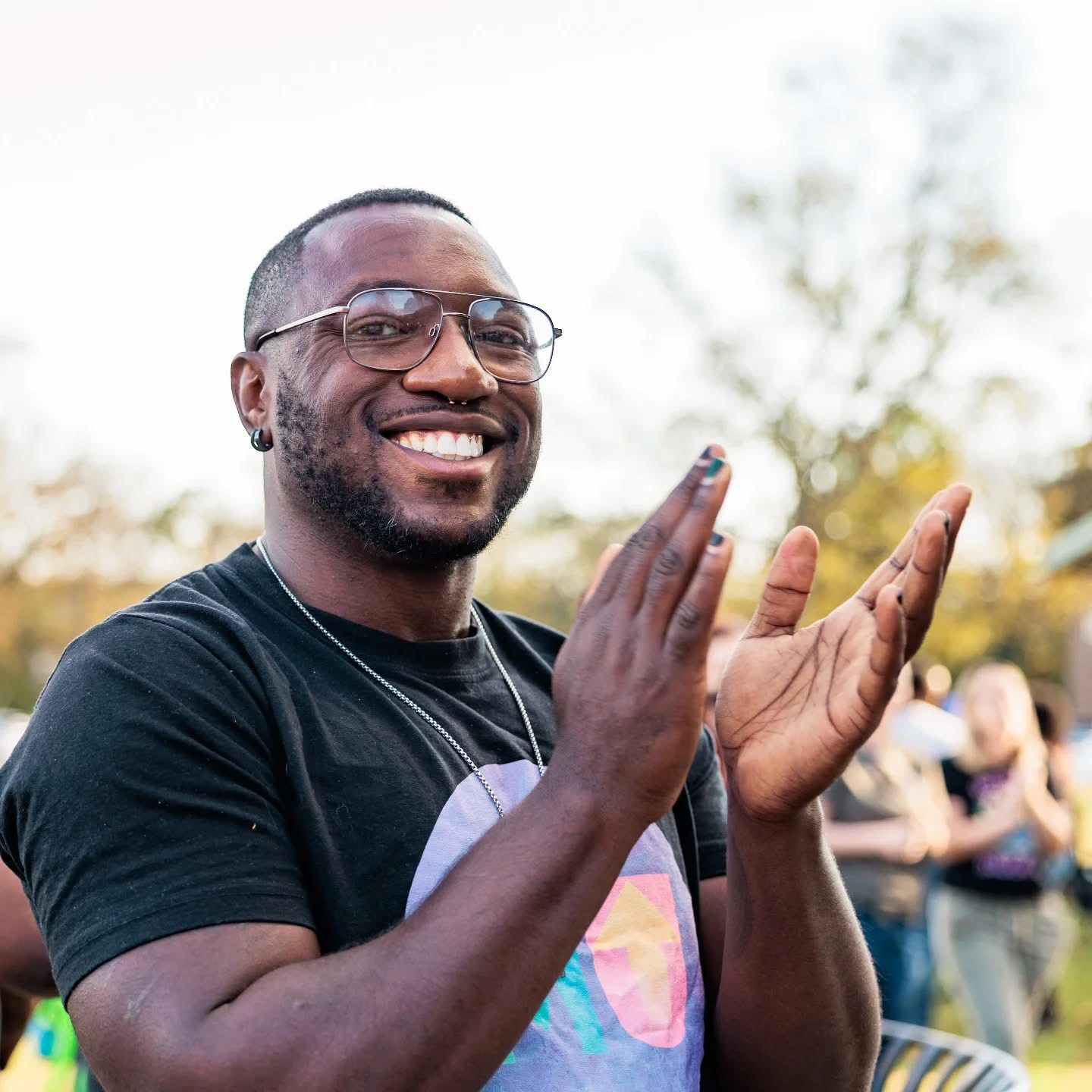 Smiling man with glasses claps outdoors, background with trees and people, sunny day.
