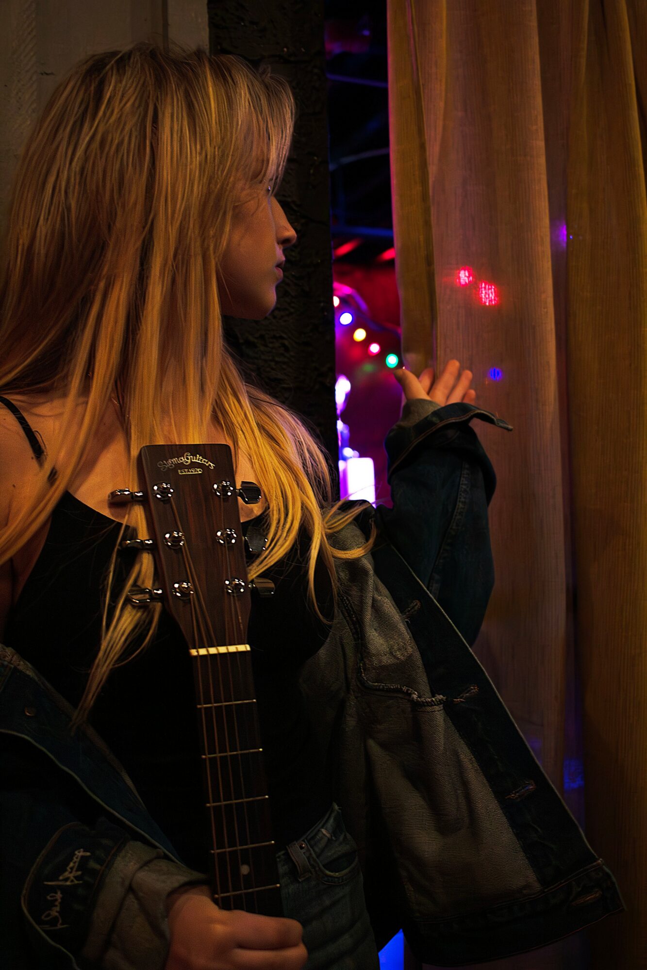 Woman with long blonde hair holding a guitar, standing near a curtain with colorful lights in the background.