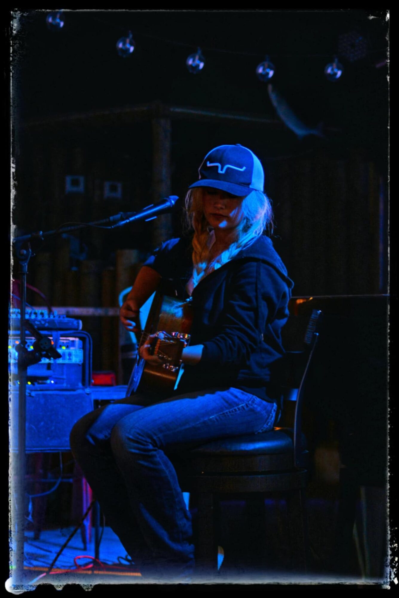 Person wearing a cap playing guitar on stage with dark background and string lights, sitting on a stool.