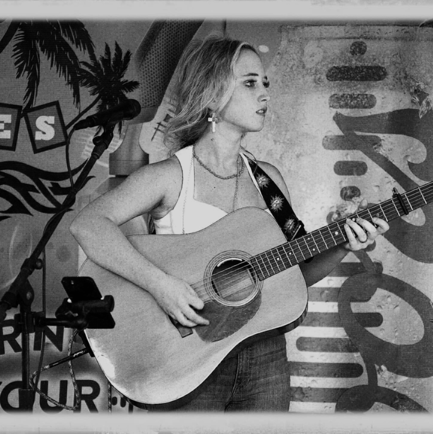 Woman playing guitar on stage with palm trees and a banner in the background, in black and white.