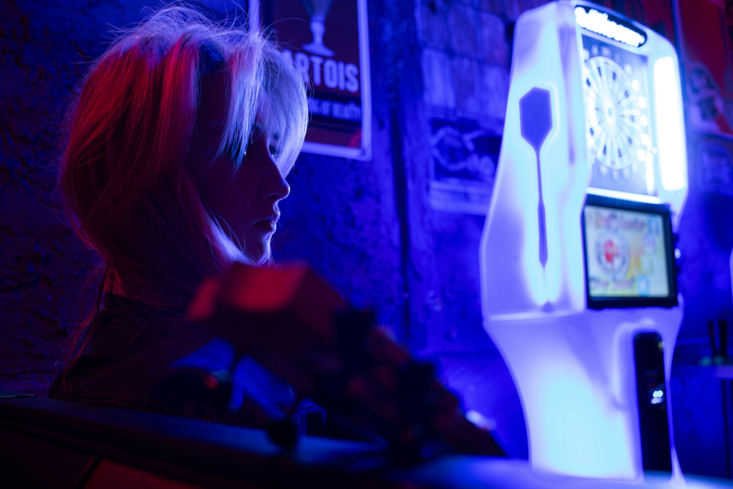 Person with light-colored hair in a dark room illuminated by blue and red lights, near a gaming machine.