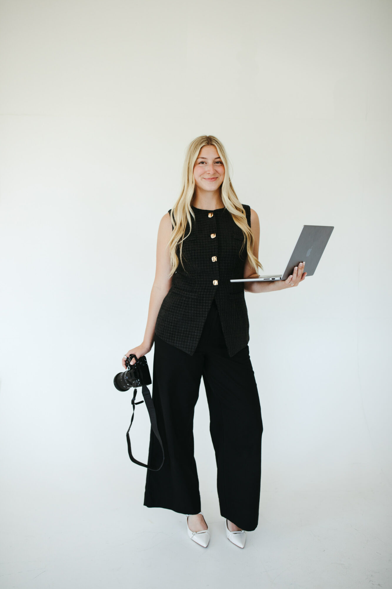 Woman with long blonde hair holding a camera in one hand and a laptop in the other, standing against a plain white background.