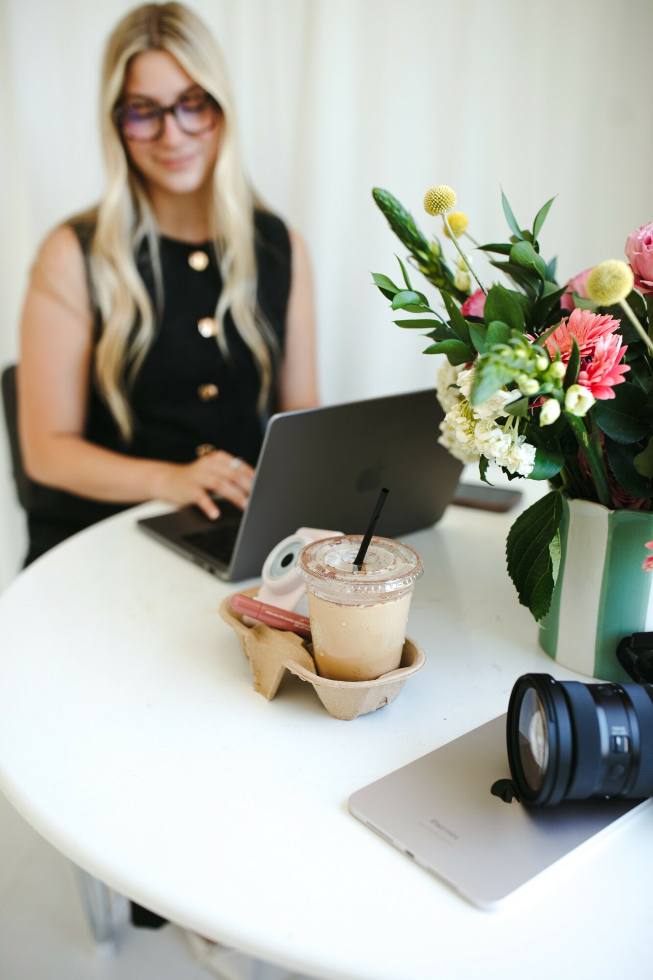 Woman with long blonde hair and glasses working on a laptop at a white table with a flower arrangement and a camera.