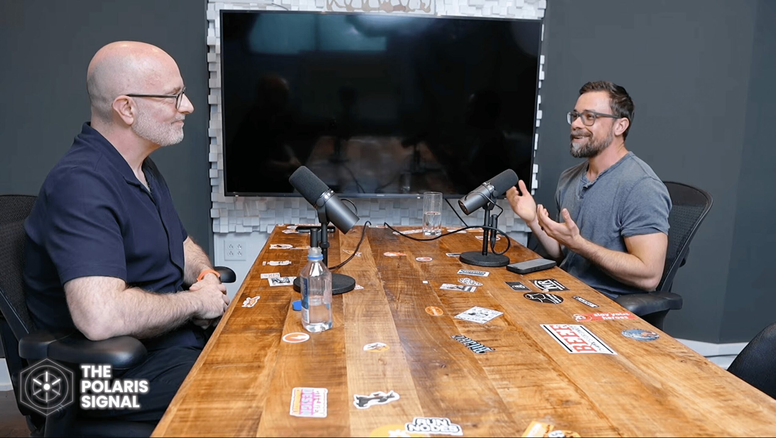 Two men sit at a wooden table with microphones, engaging in conversation, with a large screen behind them.