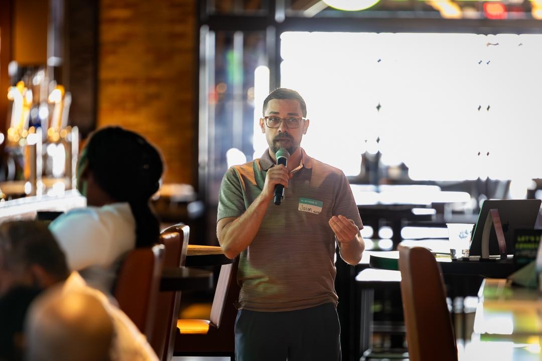 Person speaking into microphone in a restaurant or cafe, with people seated around, backlit by large window.