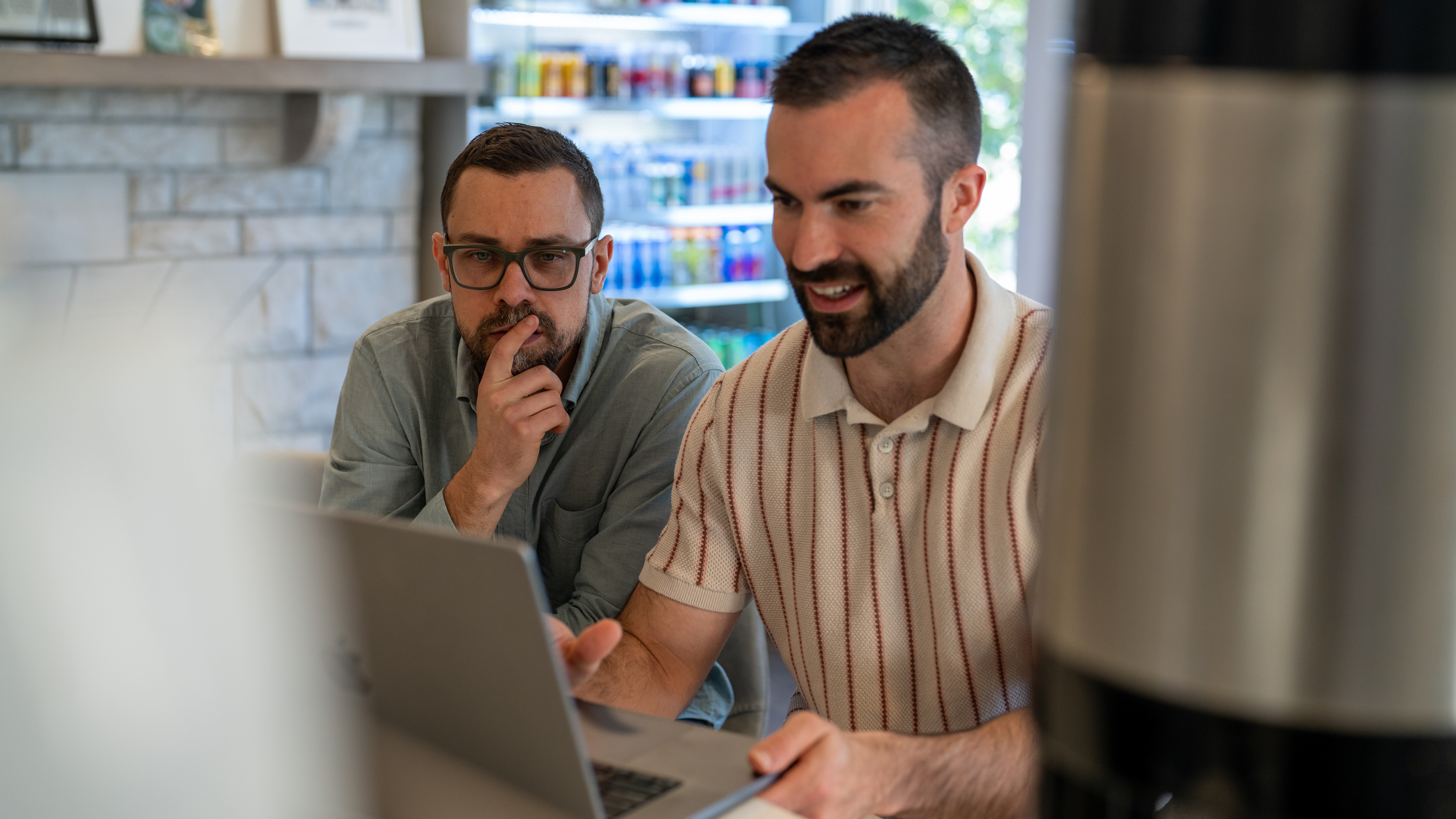 Two men sit at a table with a laptop, one smiling and the other thinking, in a bright indoor setting.