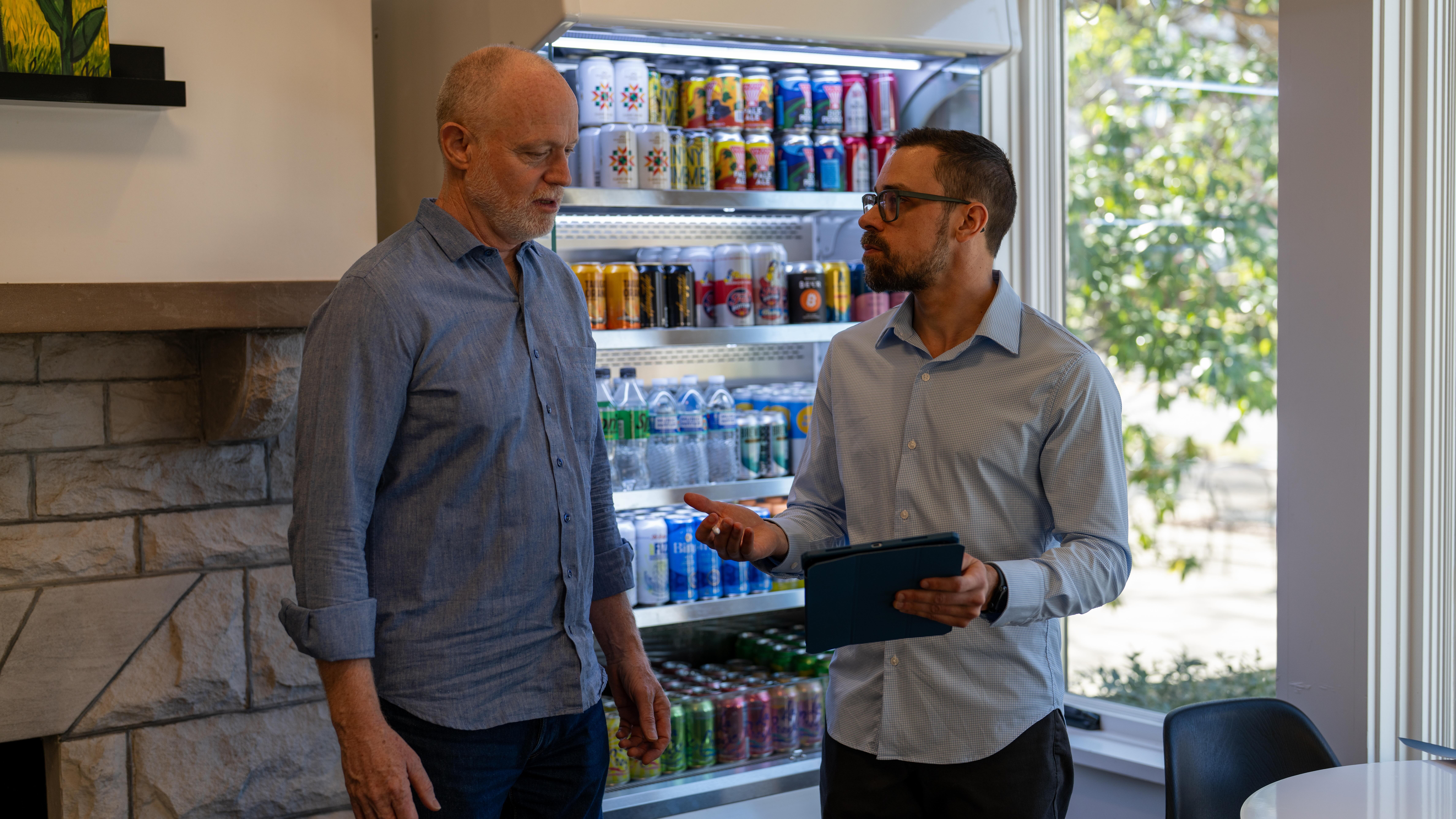 Two men are standing and talking near a vending machine filled with drinks, inside a room with a window showing trees outside.