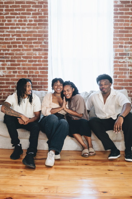 Four people sitting on a bench in front of a brick wall and window, smiling and relaxing together.