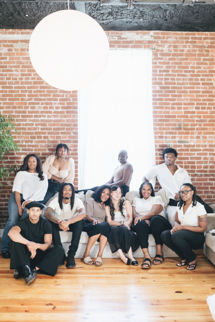 Group of nine people sitting and standing in front of a large window with brick wall background.