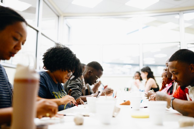 Group of people sitting at a table in a bright room, eating and talking, with large windows in the background.